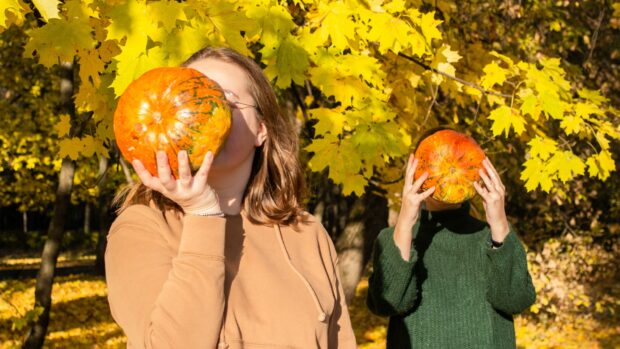 Två unga kvinnor håller pumpor framför sina ansikten, en närmare och den andra längre bort, skämtar omkring. Höstlovsroligheter under höstsäsongen